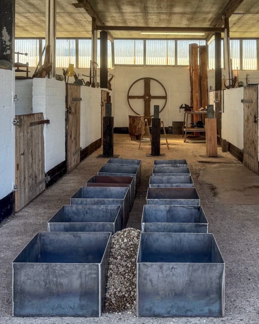 Blick durch den Werkstattgang mit symmetrisch aufgereihten Metallkästen für Schüttobjekte, im Hintergrund Skulpturen und ein altes Wagenrad an der Wand.